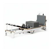 Pilates reformer machine with a white metal frame, natural wood accents, gray padded foam block, and gray backrest, shown on a clean white background.